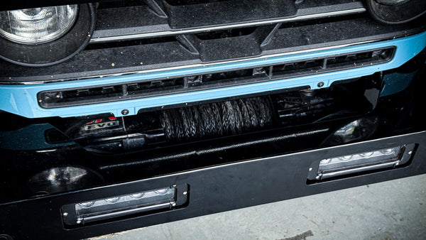 Close-up of the integrated winch mount on the INEOS Grenadier bumper, showing the synthetic winch line, fairlead, and auxiliary LED fog light housing.