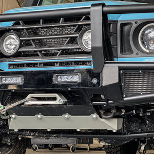 Blue INEOS Grenadier with modular front bumper system featuring a bullbar, integrated winch mount, and heavy-duty skid plate in a workshop.