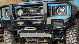 Blue INEOS Grenadier with modular front bumper system featuring a bullbar, integrated winch mount, and heavy-duty skid plate in a workshop.
