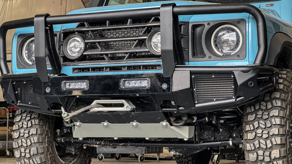 Blue INEOS Grenadier with modular front bumper system featuring a bullbar, integrated winch mount, and heavy-duty skid plate in a workshop.