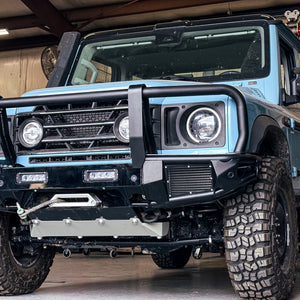 Close-up of an INEOS Grenadier featuring a modular front bumper system with an integrated 13,500 lb. winch mount, heavy-duty bullbar, and aluminum skid plate in a shop setting.