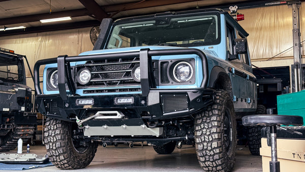 Close-up of an INEOS Grenadier featuring a modular front bumper system with an integrated 13,500 lb. winch mount, heavy-duty bullbar, and aluminum skid plate in a shop setting.